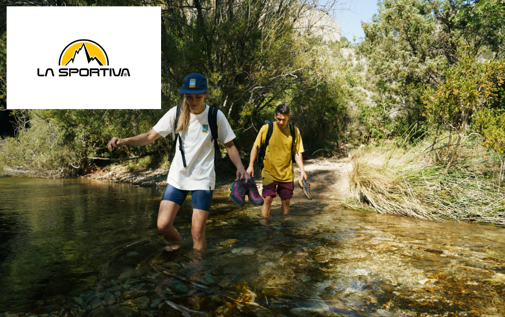 Zwei Leute überqueren einen Fluss mit ihren La Sportiva Approach Schuhen in der Hand und Outdoor Equipment von La Sportiva